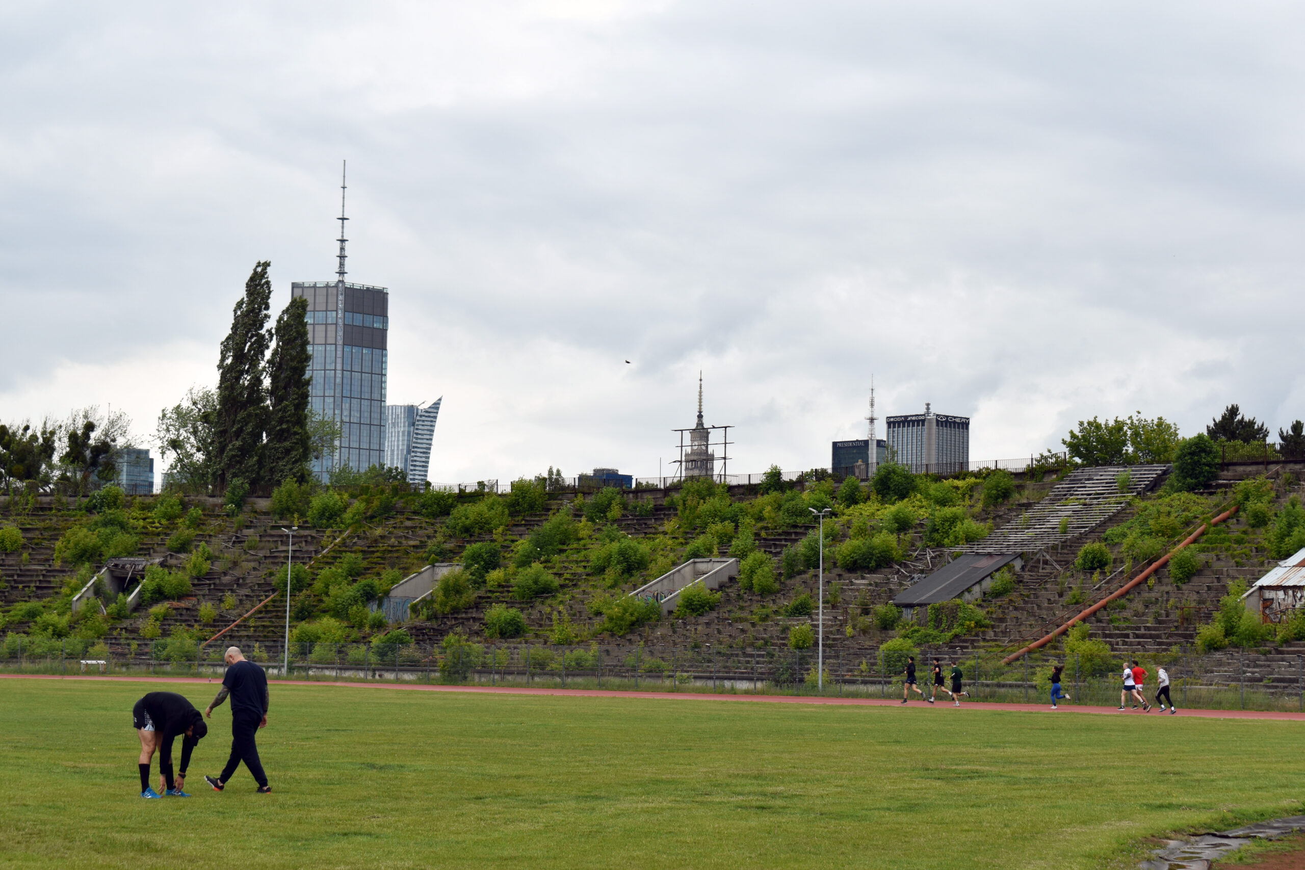 Sportowcy trenują na stadione na tle trybun zarośniętych roślinnością. Z tyłu widać panoramę Warszawy.