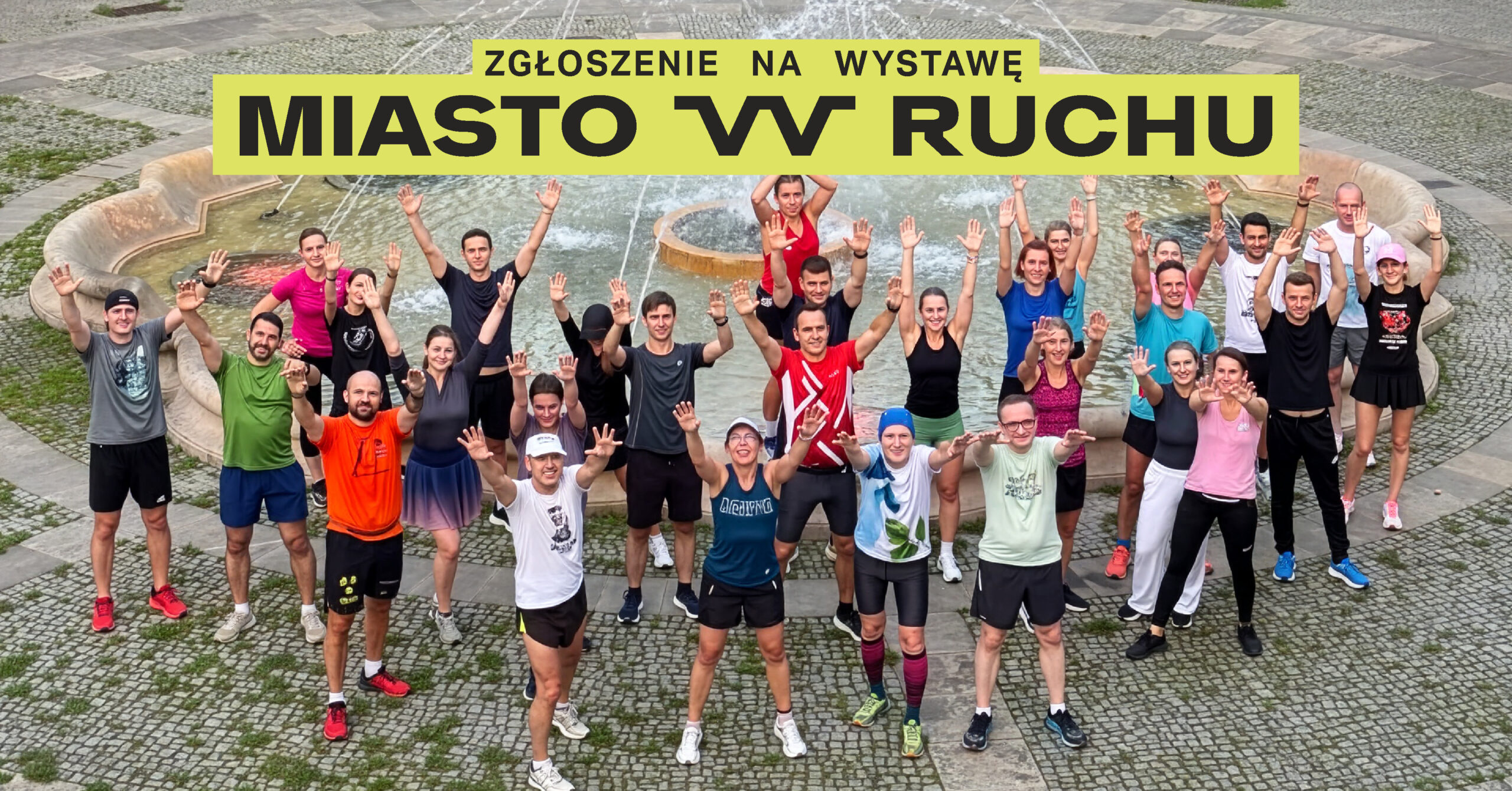 Grupa ludzi w kolorowych, sportowych ubraniach podnosi ręce do góry na tle fontanny miejskiej.